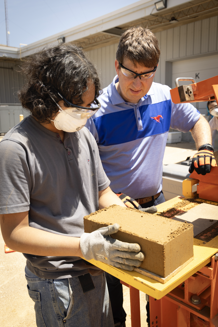 A teacher and a student stand holding a newly compressed earth block