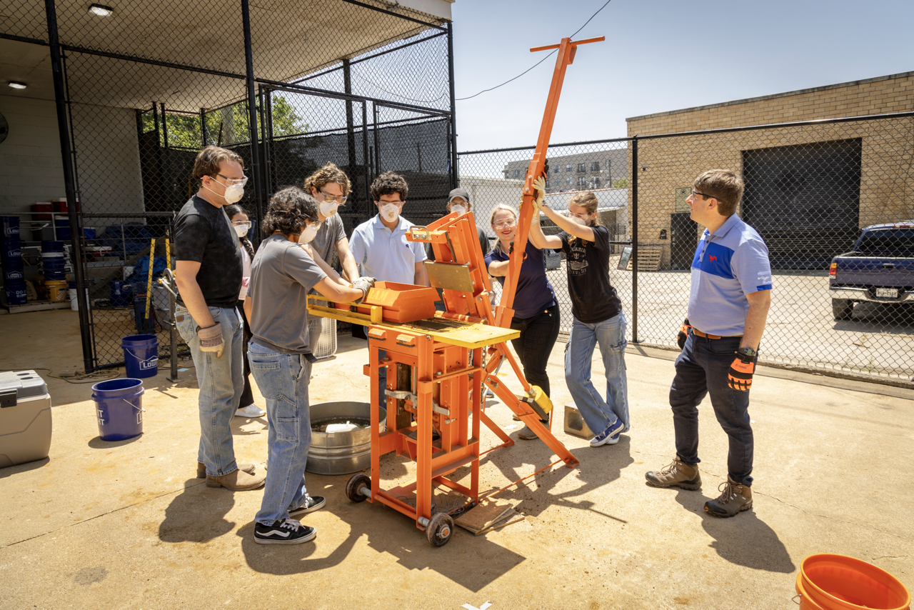 A group of students surround a machine used to compress and mold bricks