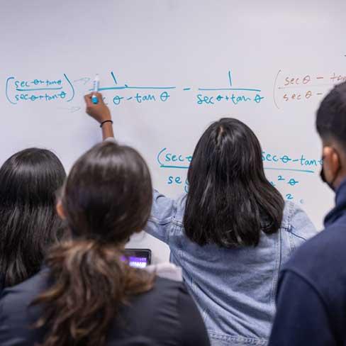 Student solving equation on whiteboard with others students gathered to observe and assist.