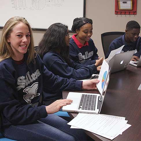 Girl in Sellers GBL program looks up from computer and business graph to smile at camera while students nearby study documents.
