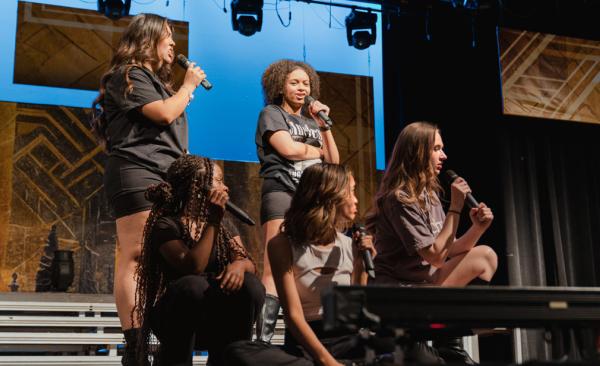 A closeup shot during a performance of "Six" with several students singing over each other while others crouch and gaze at the singers.