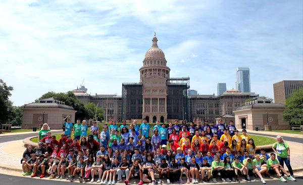 A large group of students sit, kneel, and stand in front of the Texas Capitol building.