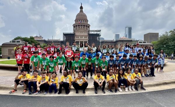 A large group of students sit, kneel, and stand in front of the Texas Capitol building.