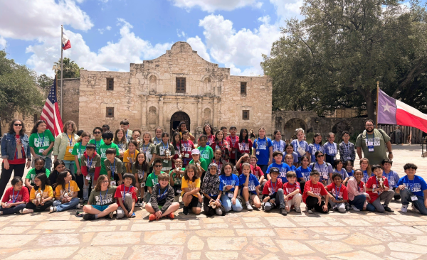 A large group of students and chaperones sit, kneel, and stand in front of the Alamo