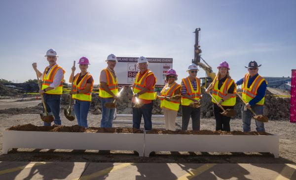 A group of adults stand in hard hats and vests with shovels extended