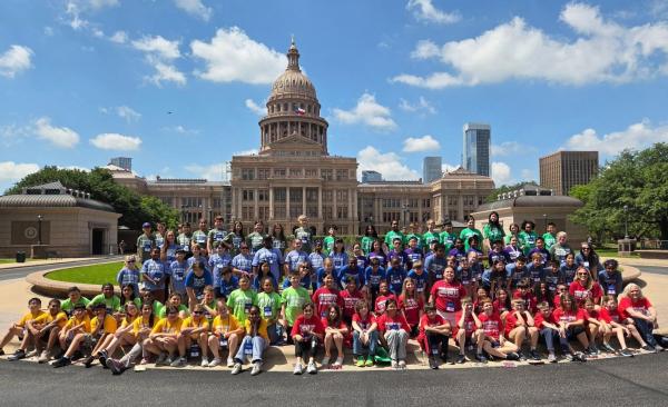 A large group of students sit, kneel, and stand in front of the Texas Capitol building.
