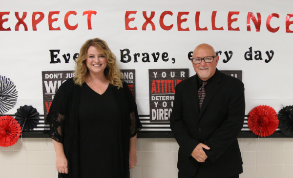 a female and a male teacher stand in front of bulletin board that reads Expect Excellence