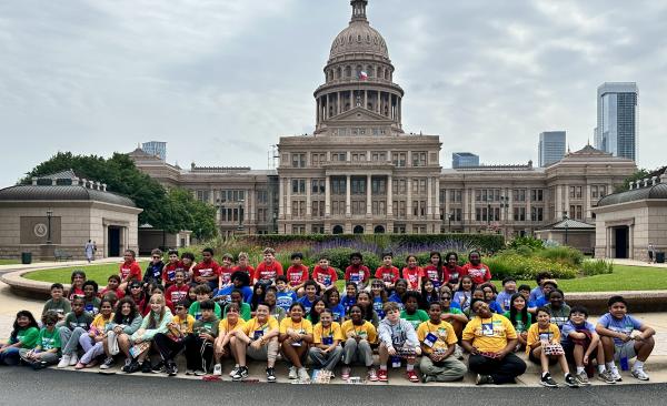 A large group of students sit, kneel, and stand in front of the Texas Capitol building.