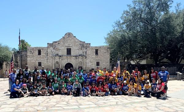 A large group of students sit, kneel, and stand in front of the Texas Capitol building.