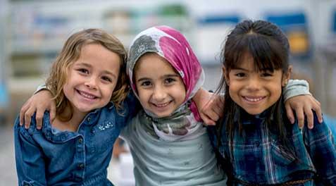 Three young girls smiling and hugging each other in a photo, one wearing a hijab.