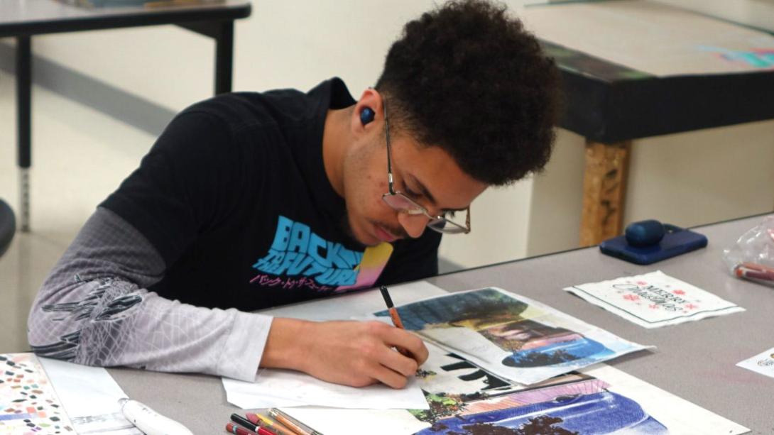 Student leans over their desk working on mixed media art work.