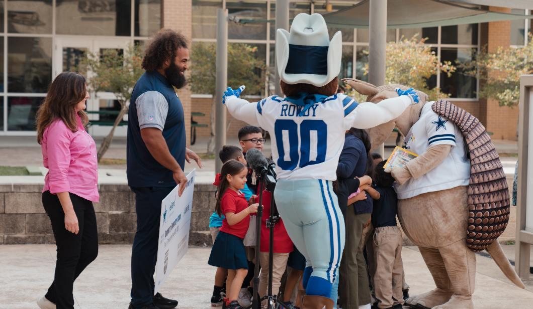  Two adults give a side hug while mascots and children applaud.