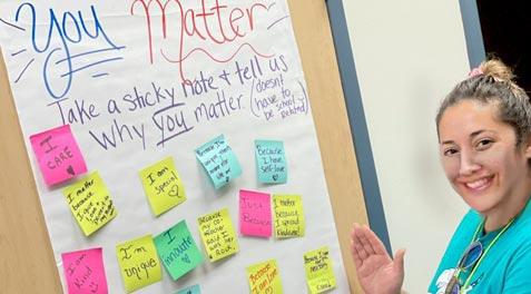 A teacher next to a poster with post it notes of students affirming their self worth.