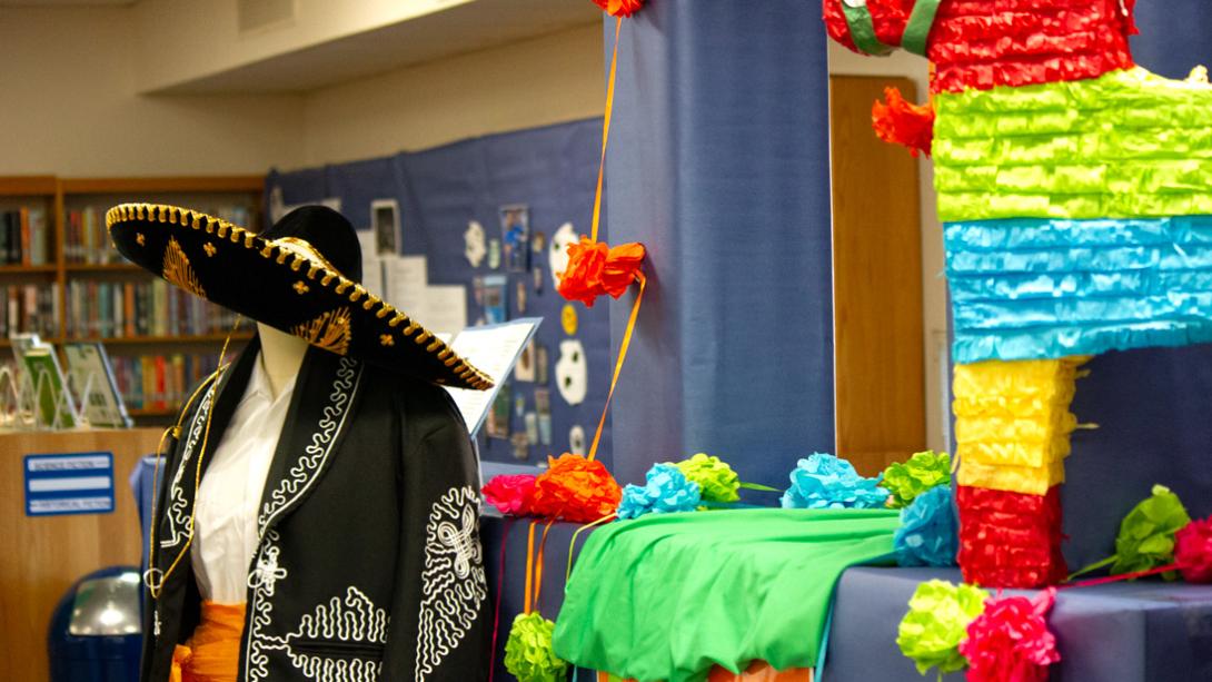 A Charro suit is displayed next to a colorful Piñata, a wall with Hispanic historical locations in the background.