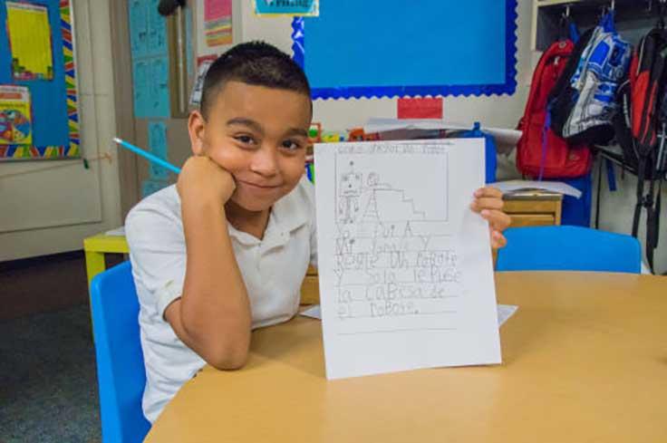 Boy with pencil in hand is showing off his paper with writing in Spanish.