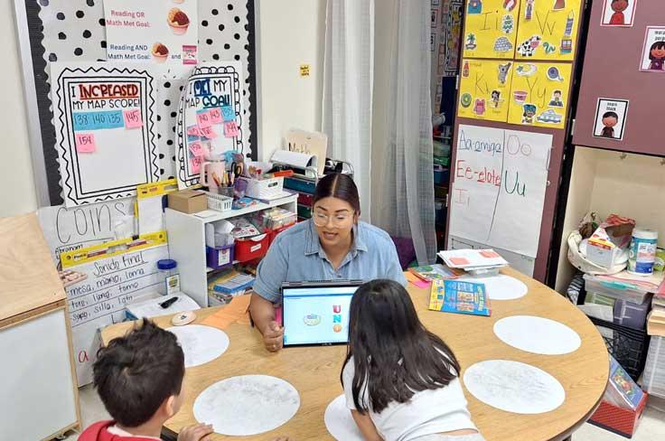Teacher is holding up an iPad showing the word "uno" while working with a couple of students.