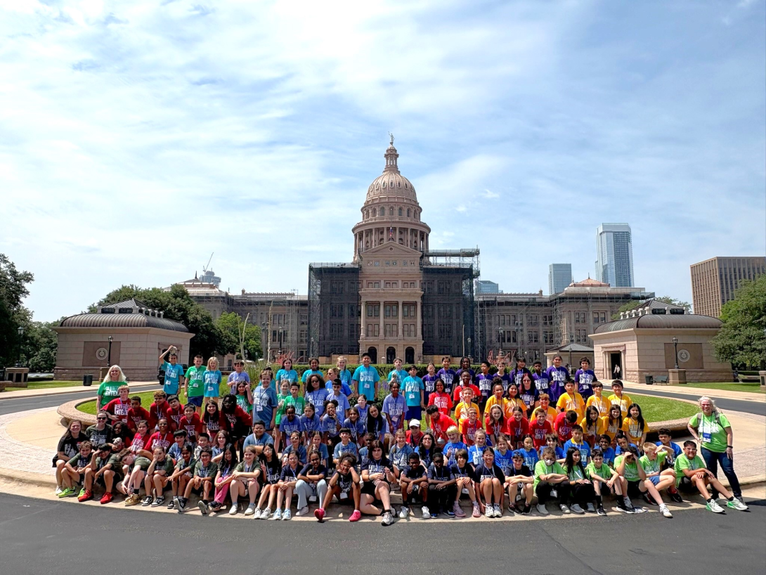 A large group of students sit, kneel, and stand in front of the Texas Capitol building.
