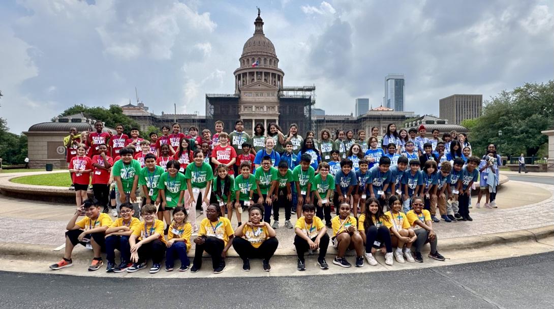 A large group of students sit, kneel, and stand in front of the Texas Capitol building.