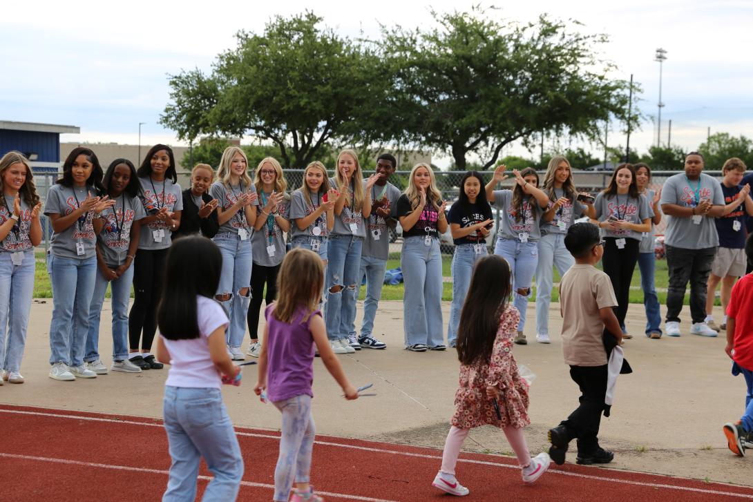 Older students stand in a line clapping while younger students walk in front of them