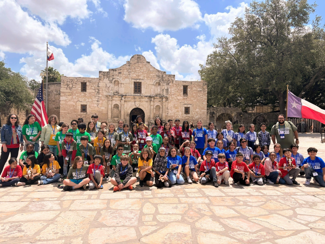 A large group of students and chaperones sit, kneel, and stand in front of the Alamo