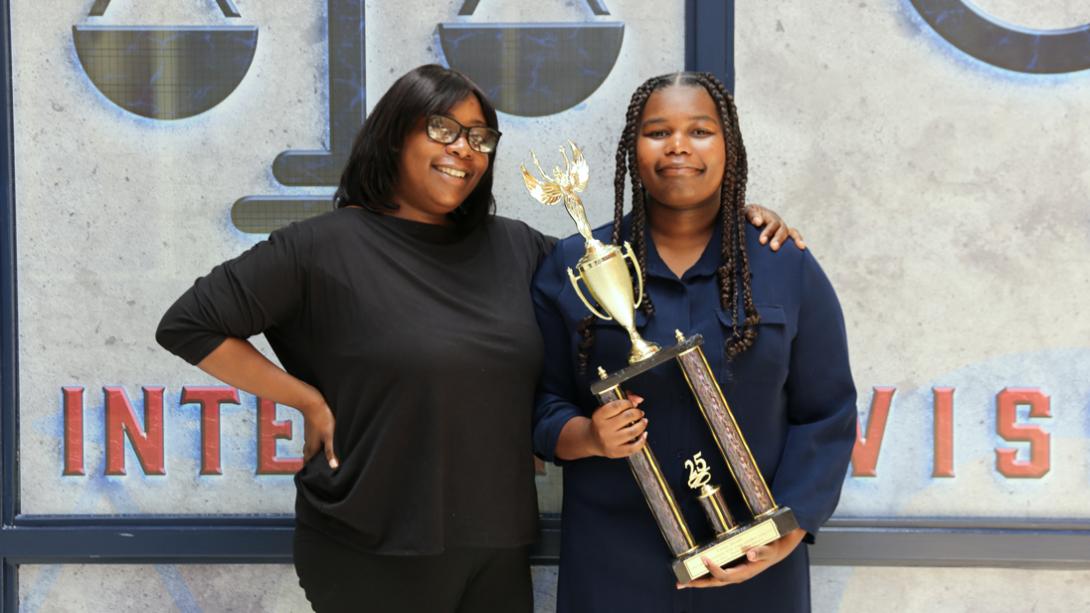 A young female student, Casey D., holds a trophy while standing together with her mom.