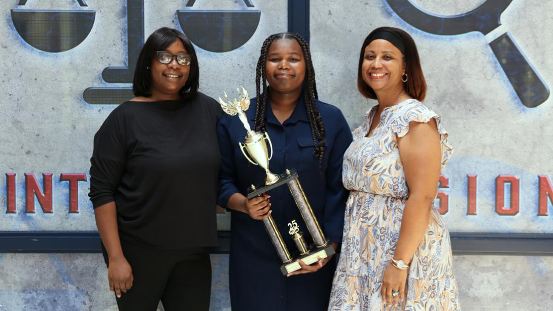 Casey poses with her former teacher and her mom while smiling and holding her trophy.