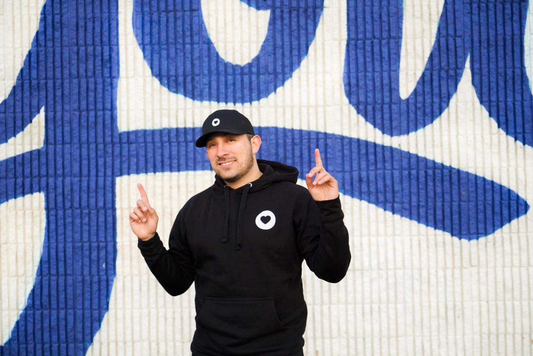 A man in a black sweatshirt and cap stands in front of a wall