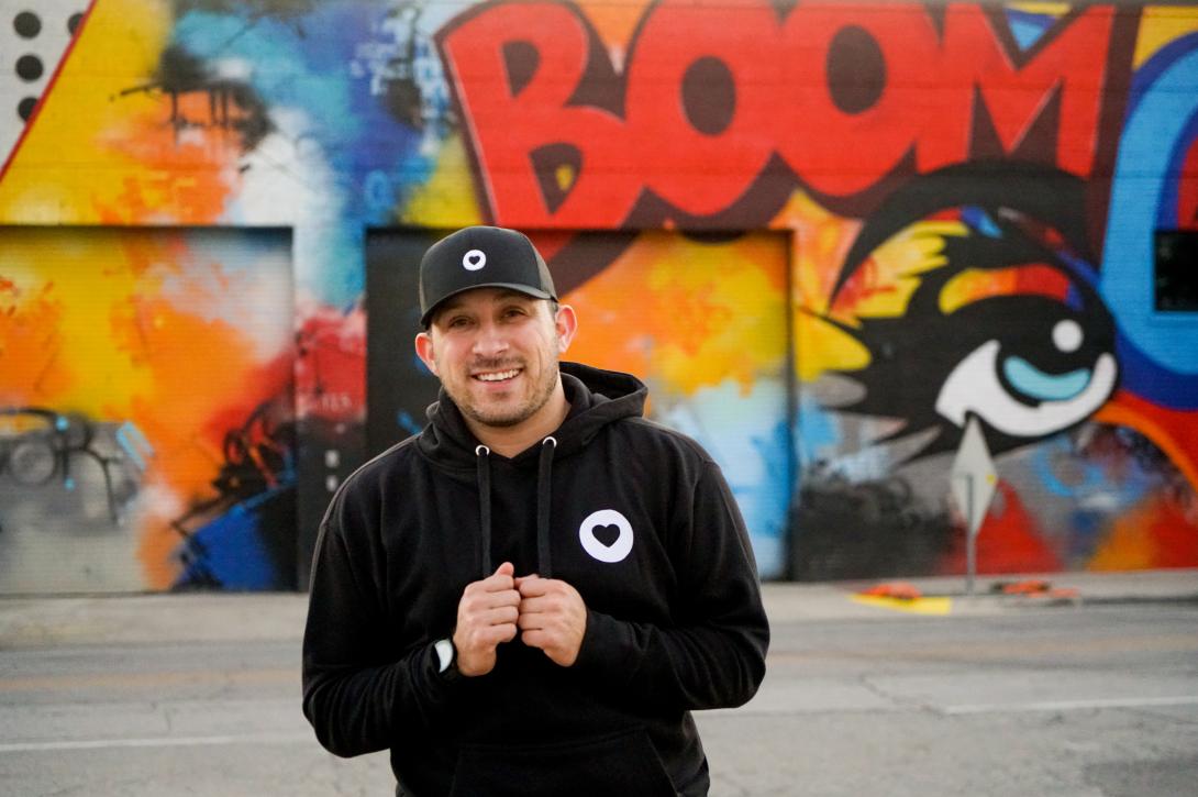 A man in a black sweatshirt and baseball cap stands smiling in front of a graffitied wall 