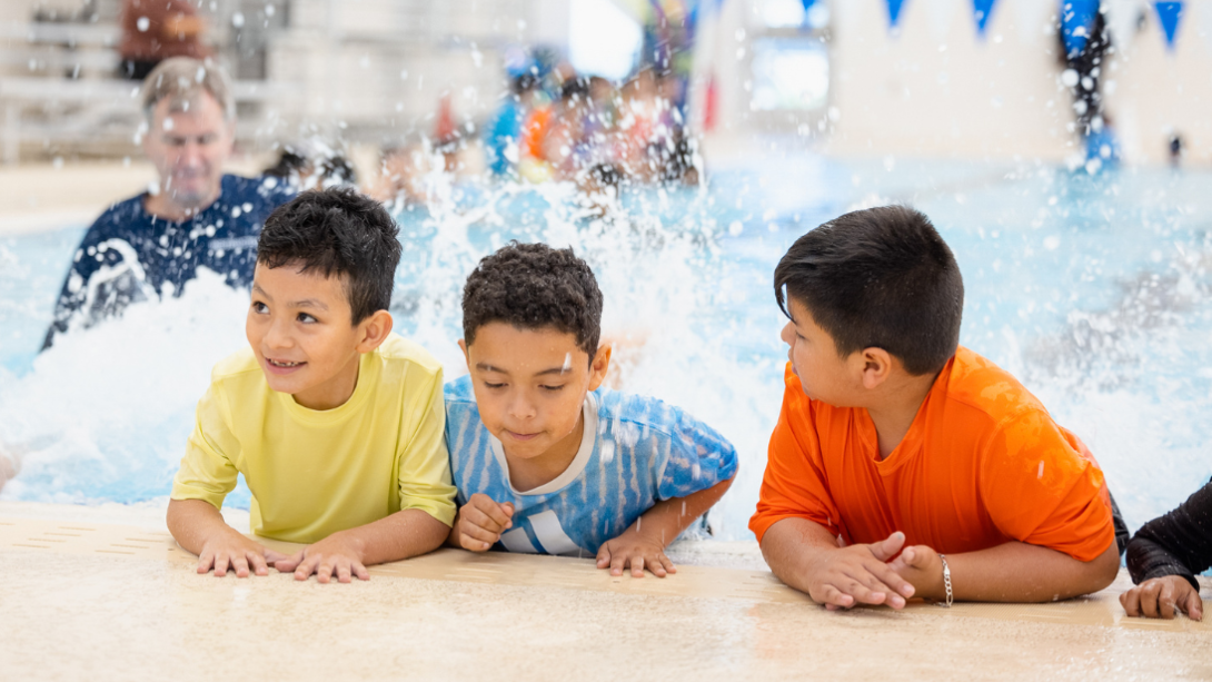three male students lean on the edge of a pool while kicking their feet in the water