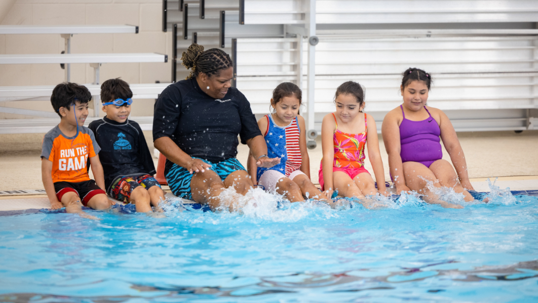 A diverse group of students and an instruction kick their feet in the water of a pool