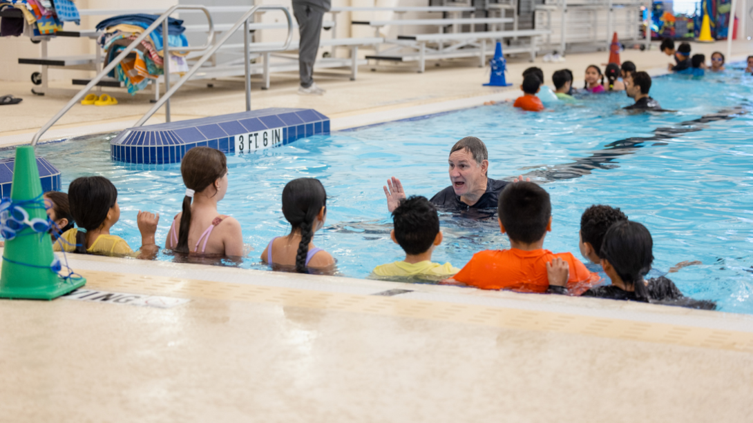 A water safety instructor speaks to a group of students in the pool