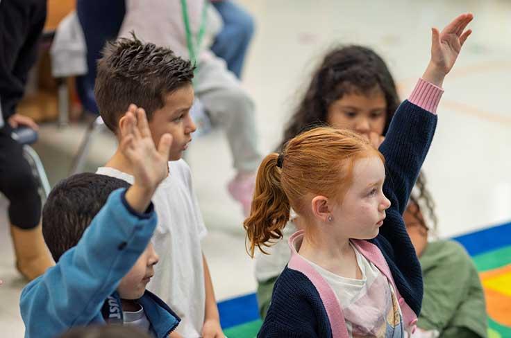 Students sitting on floor with raised hands in an early childhood classroom
