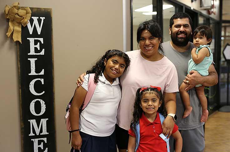 Mother and father with a baby and two school-age children standing next to a welcome sign.