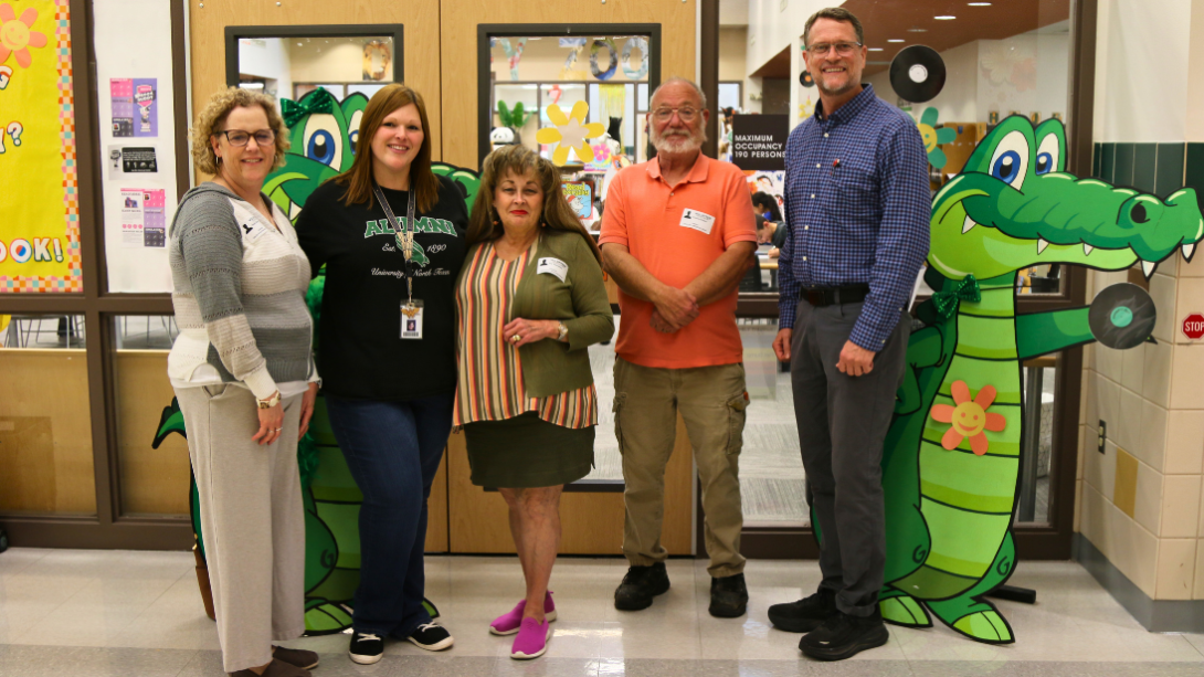 three adult women and two adult men stand together in front of an alligator cutout