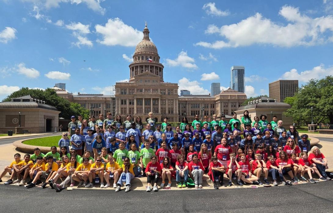A large group of students sit, kneel, and stand in front of the Texas Capitol building.