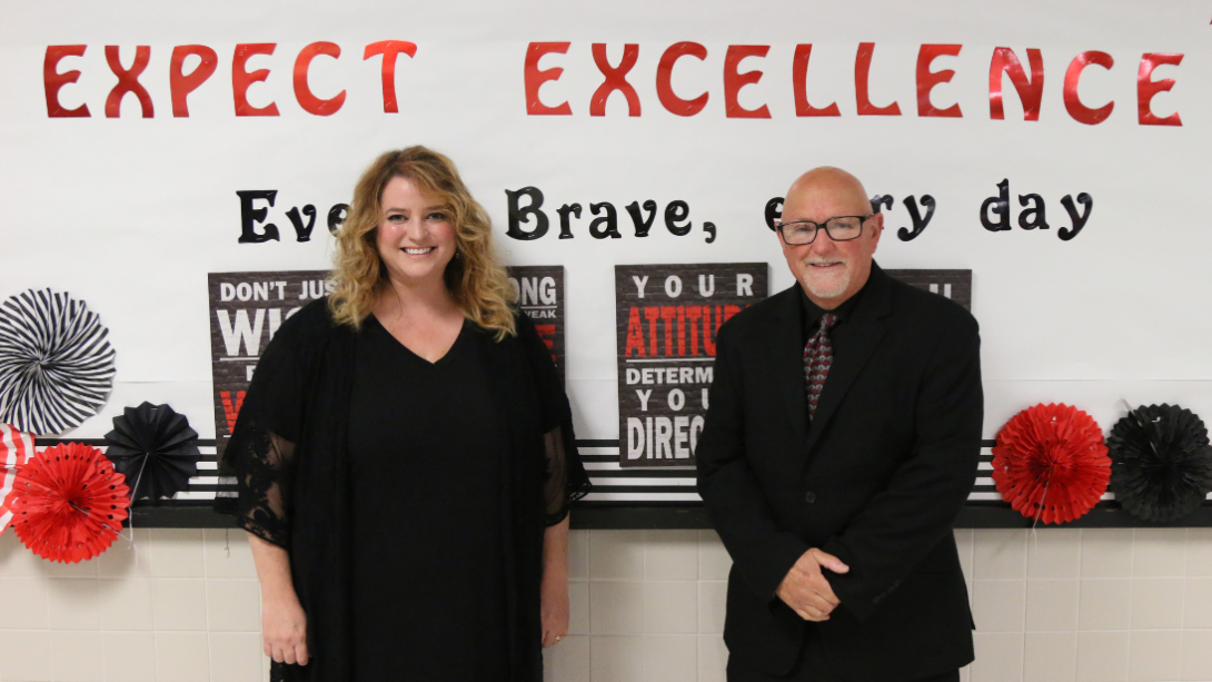 a female and a male teacher stand in front of bulletin board that reads Expect Excellence