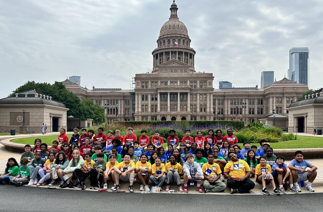 A large group of students sit, kneel, and stand in front of the Texas Capitol building.