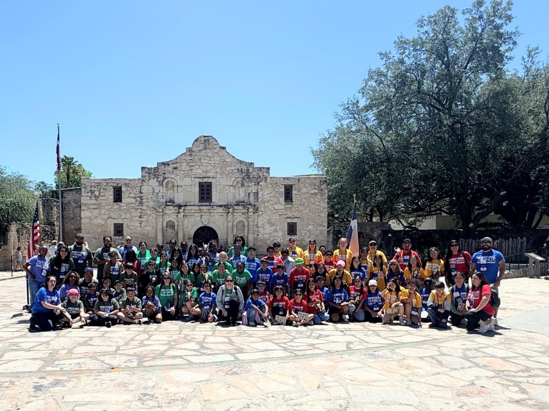 A large group of students sit, kneel, and stand in front of the Texas Capitol building.